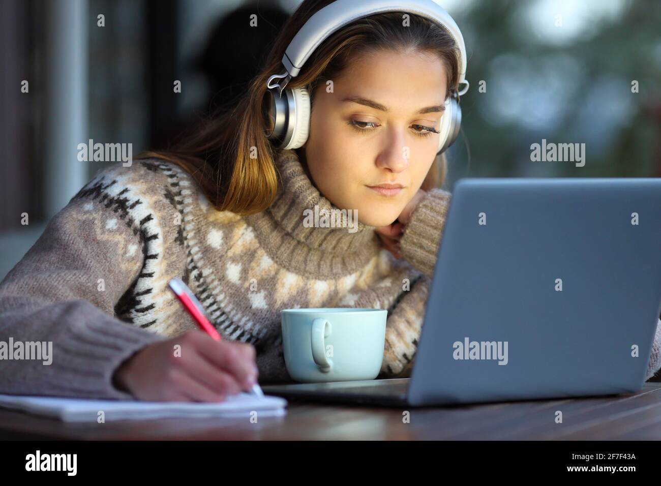 Konzentrierter Schüler mit Kopfhörern, E-Learning-Kurse und einem Laptop, der sich Notizen zu den Themen gemacht hat Notebook im Winter in einem Café Stockfoto