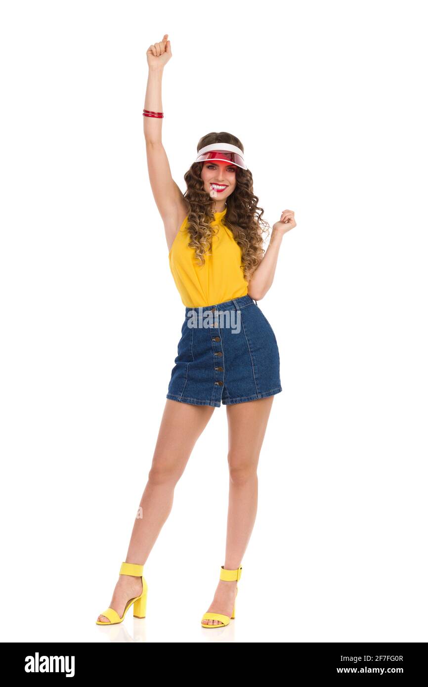 Glückliche junge Frau in High Heels Sandalen, Jeans Minirock, gelbes Oberteil und rotem Sonnenschirm steht mit erhobenem Arm und lächelnd. Vorderansicht. Volle Leng Stockfoto