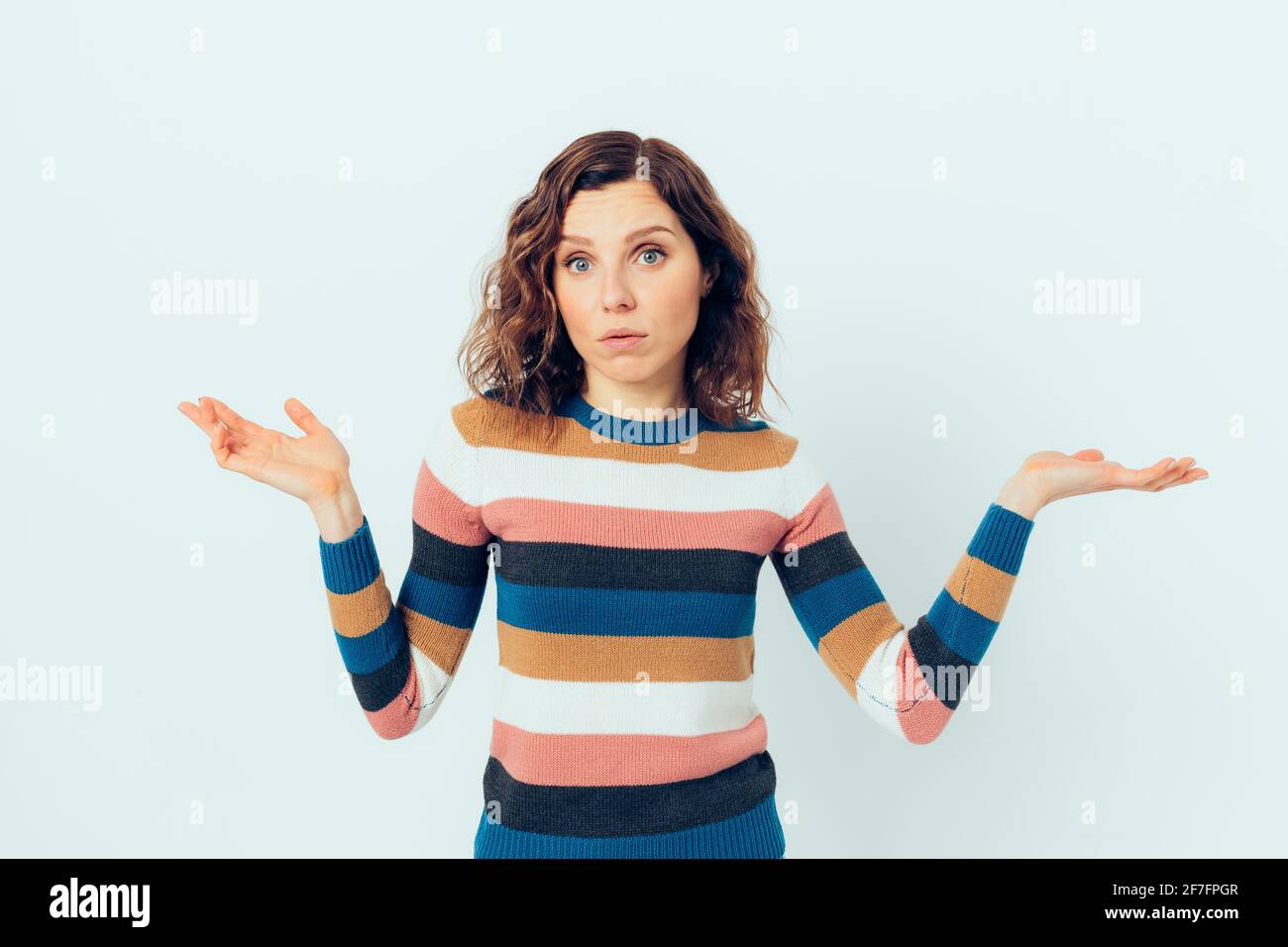 Die junge Frau, die einen lässigen gestreiften Pullover trägt, zuckt mit gleichgültigem Ausdruck mit den Schultern, während sie vor weißem Hintergrund steht. Stockfoto