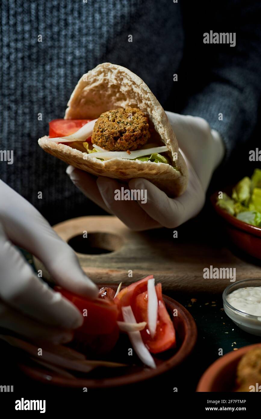 Ein junger Mann, der Latexhandschuhe trägt, bereitet ein Falafel-Sandwich in einem Pita-Brot mit gehackten Tomaten und Zwiebeln zu Stockfoto