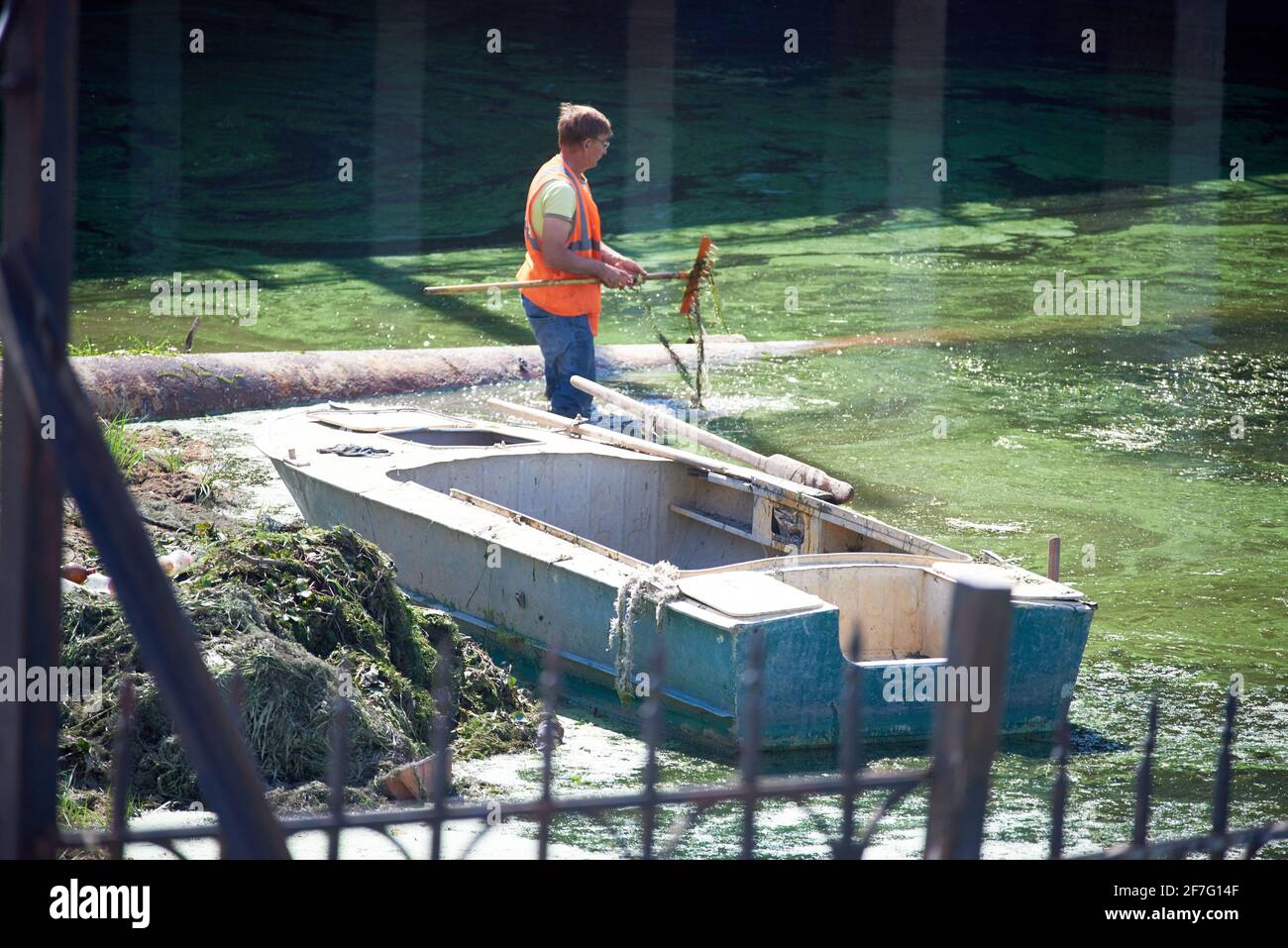 Ein Mann in einer orangefarbenen Signalweste räumt Schlamm aus dem Fluss. Es gibt ein Boot in der Nähe. Stockfoto