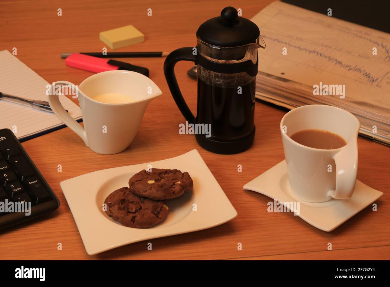 Kaffeetasse und Krug mit Milch, machen Sie eine Pause, trinken Sie und zwei Schokoladenkekse auf dem Teller, Dokumente, Tastatur, Stift, Stift und Pad Stockfoto
