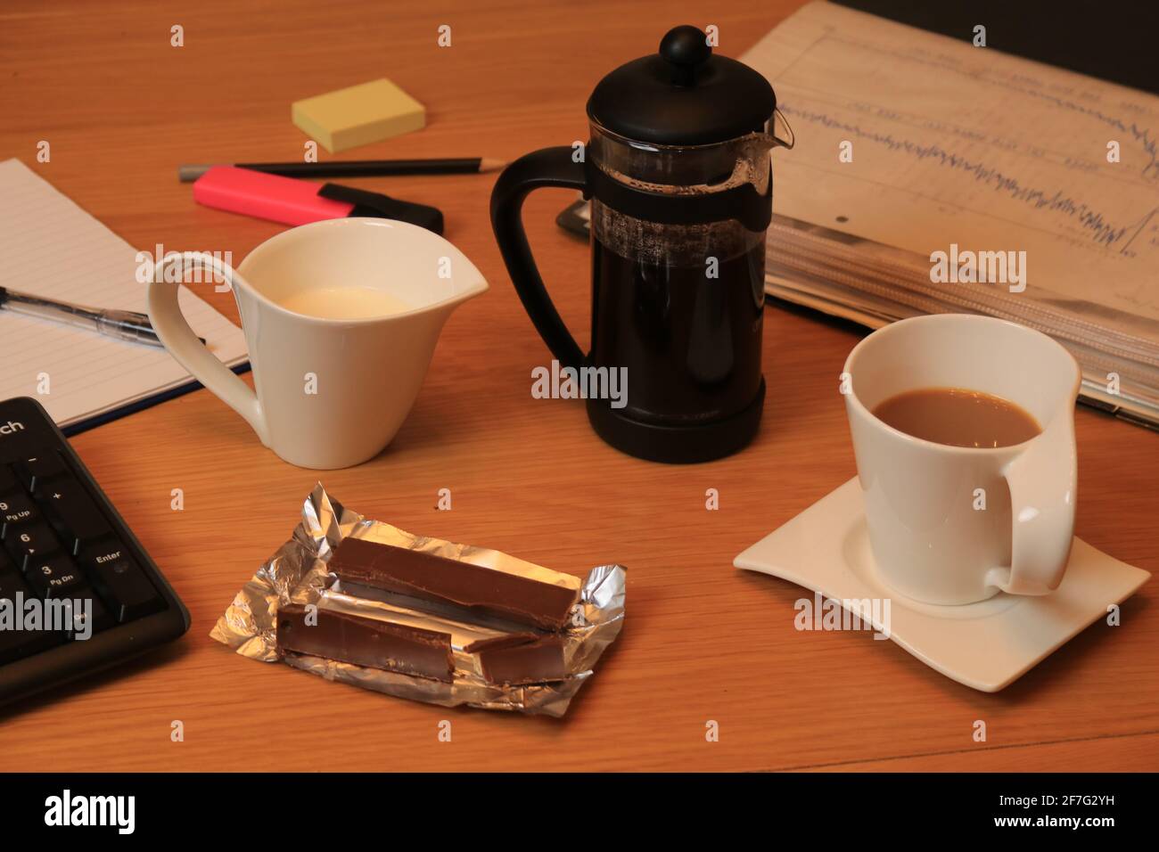 Kaffeetasse und Kanne mit Milch, machen Sie eine Pause, trinken und mit Wafer-Schokolade überzogene Snacks in Zinnfolie, Dokumenten, Tastatur, Stift, Bleistift und Pad Stockfoto