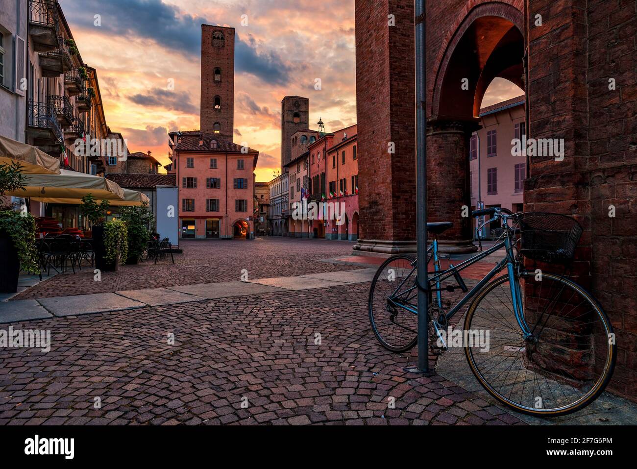 Fahrrad auf dem gepflasterten Stadtplatz, umgeben von alten Häusern und mittelalterlichen Türmen, unter dem wunderschönen Abendhimmel in Alba, Piemont, Norditalien. Stockfoto