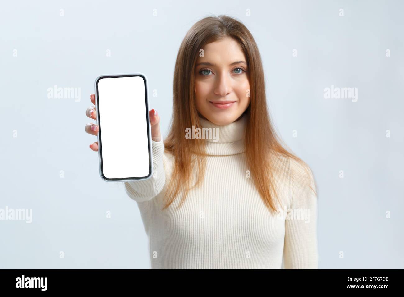 Lächelnde junge Frau zeigt Smartphone mit weißem Touchscreen-Mockup. Schöne braune haarige Mädchen auf einem grauen Hintergrund. Stockfoto