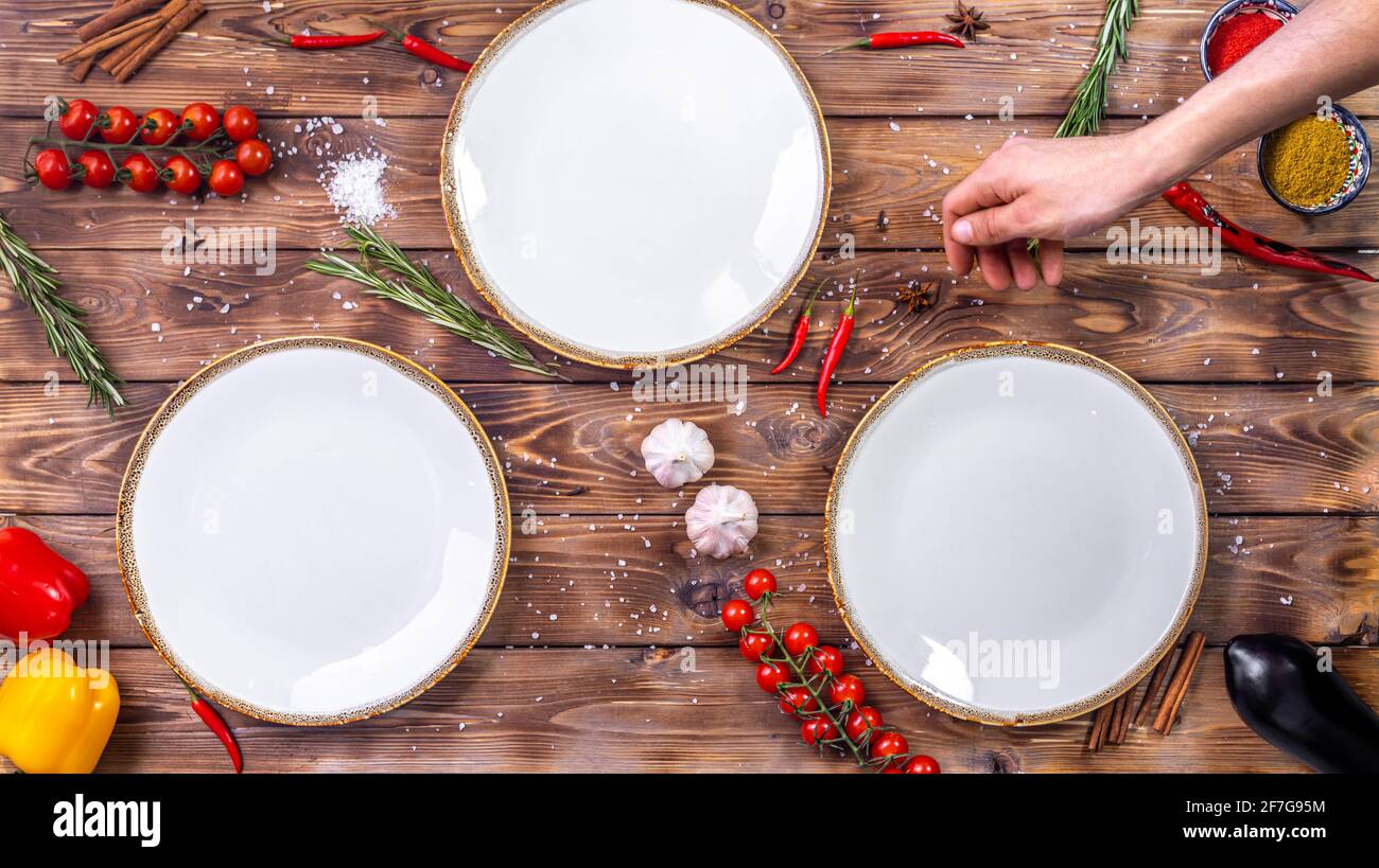 Leere Teller stehen auf einem braunen Holzhintergrund, und der Food Stylist schmückt sie mit Kirschtomaten, Rosmarin, Chilli und grobem Salz. Fl Stockfoto