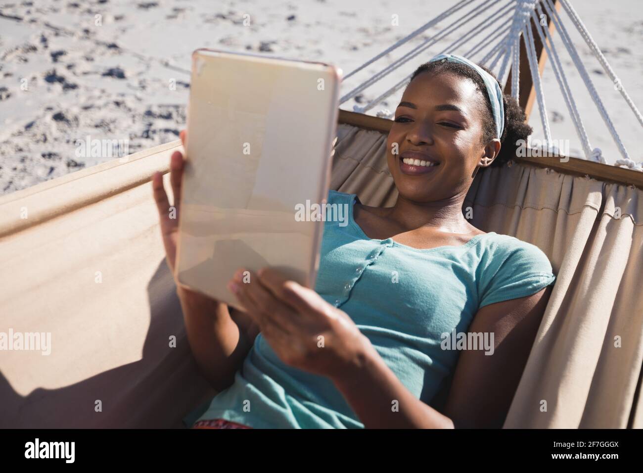 Glückliche afroamerikanische Frau, die in der Hängematte am Strand liegend benutzt Tablet Stockfoto