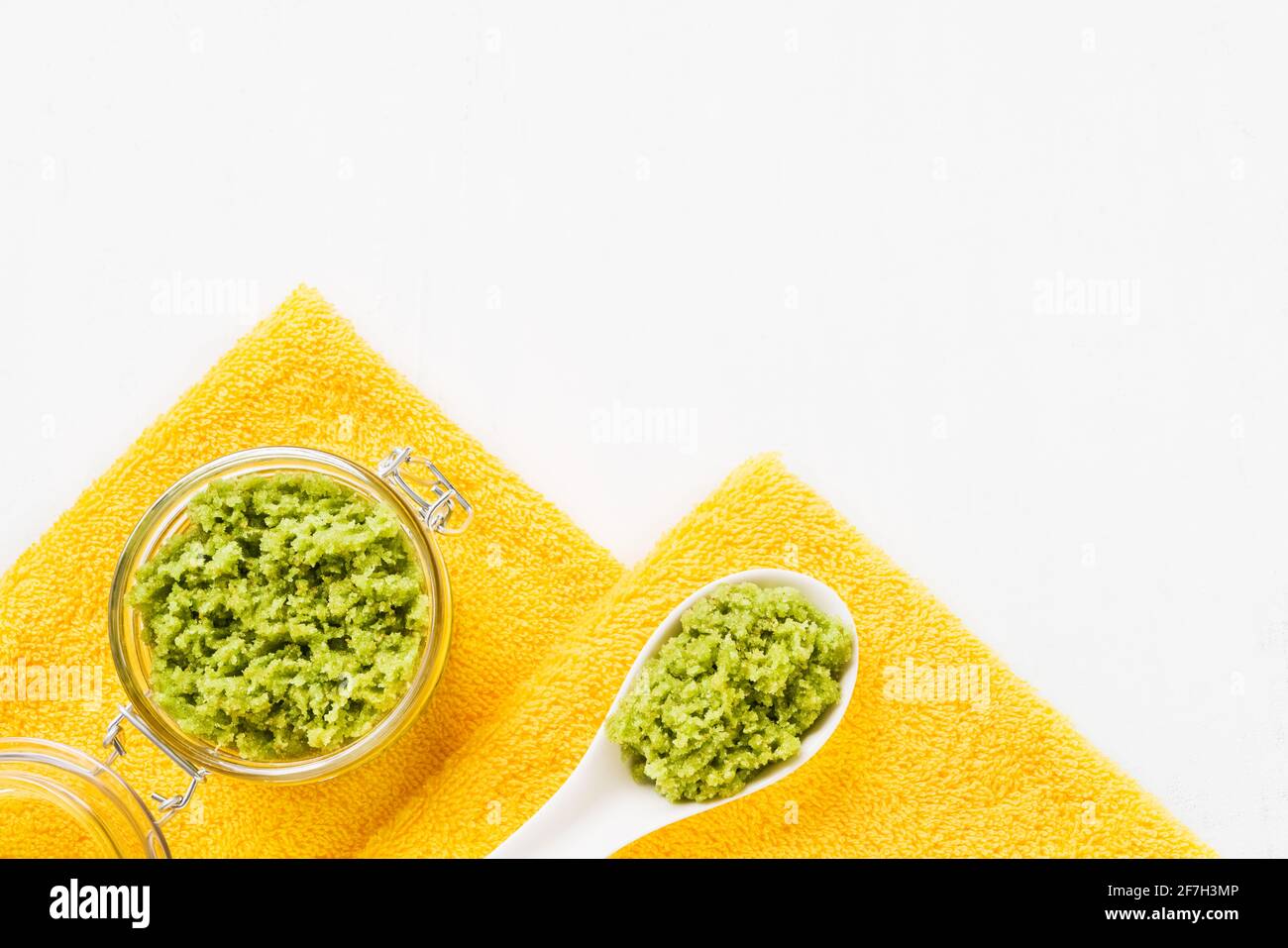 Grüner Körper schrubbt und gefaltete gelbe Handtücher auf einem weißen Holzhintergrund. SPA, Hygiene, Wellness-Wohlbefinden, Körperpflege-Konzept. Platz zum Kopieren, flach liegend Stockfoto