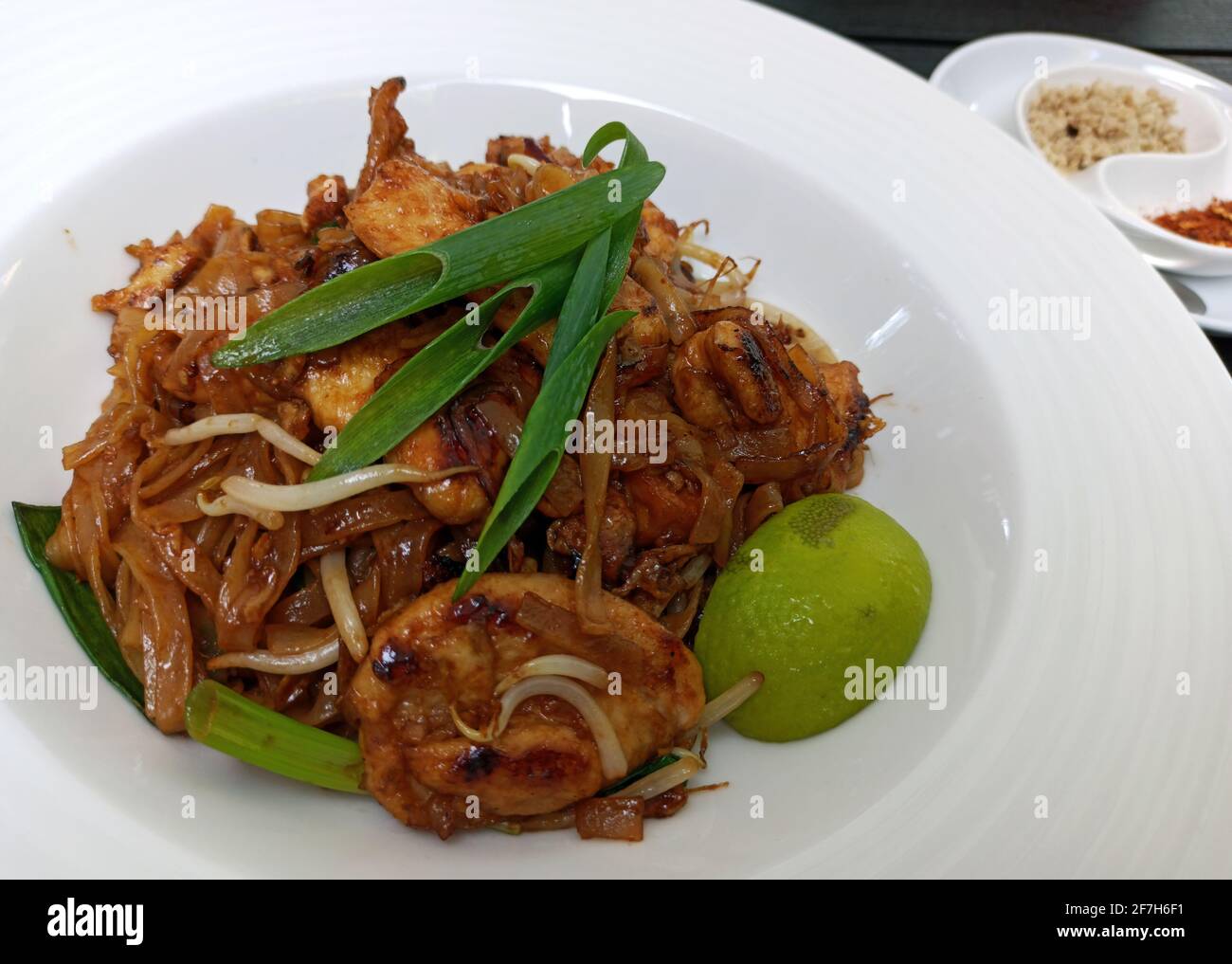 Pad thai Nudeln mit gebratenem Huhn, gemischte grüne Blätter, gehackte Erdnüsse, Schalotten & Limettenkeil auf weißem Teller vor Holzhintergrund serviert heiß wi Stockfoto