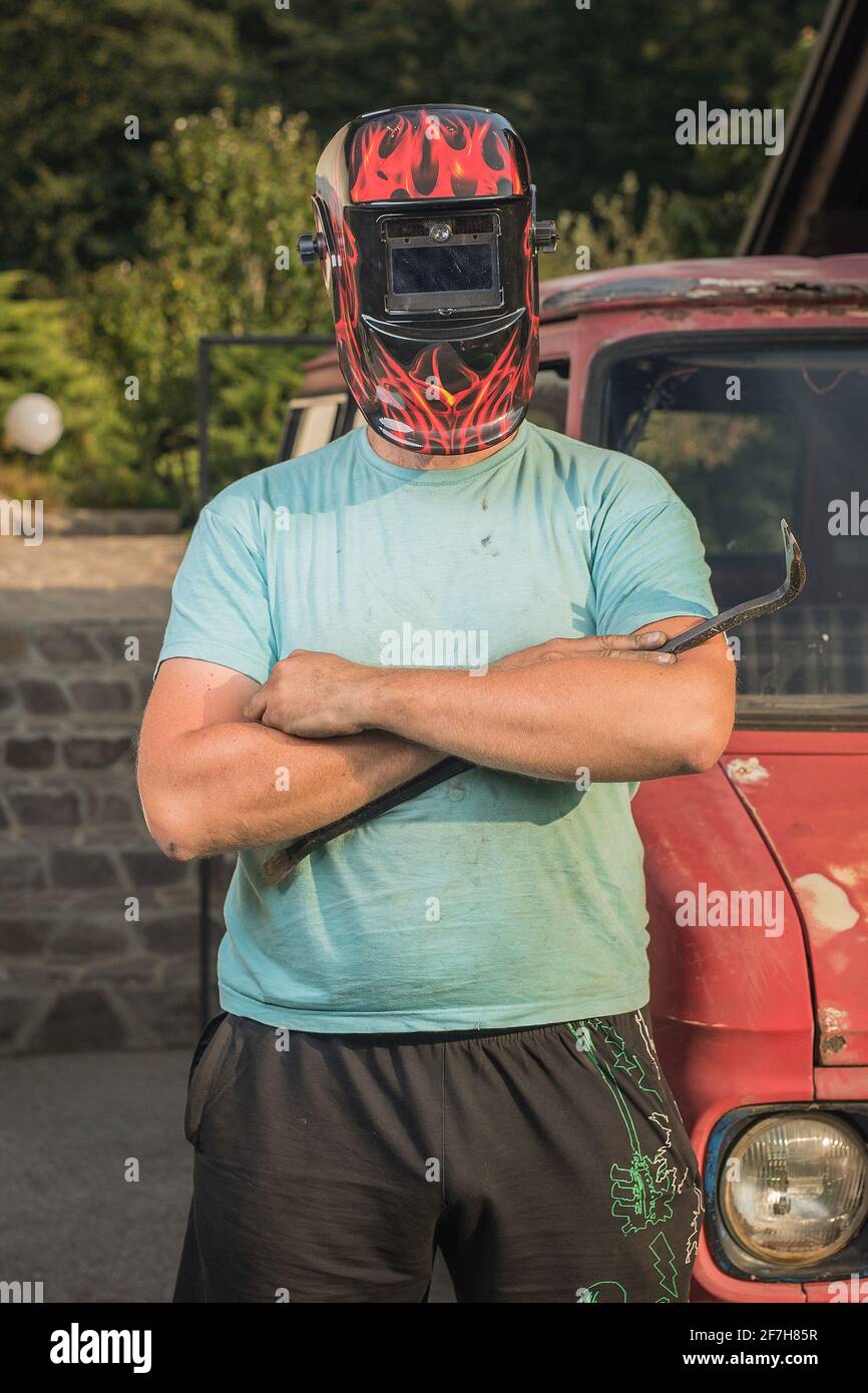 Mann mit Schweißmaske auf dem Kopf vor einem rostigen und zerstörten Oldtimer-LKW oder -Van. Auto wird wiederhergestellt. Harter Kerl, der ein Auto wieder herstellt Stockfoto