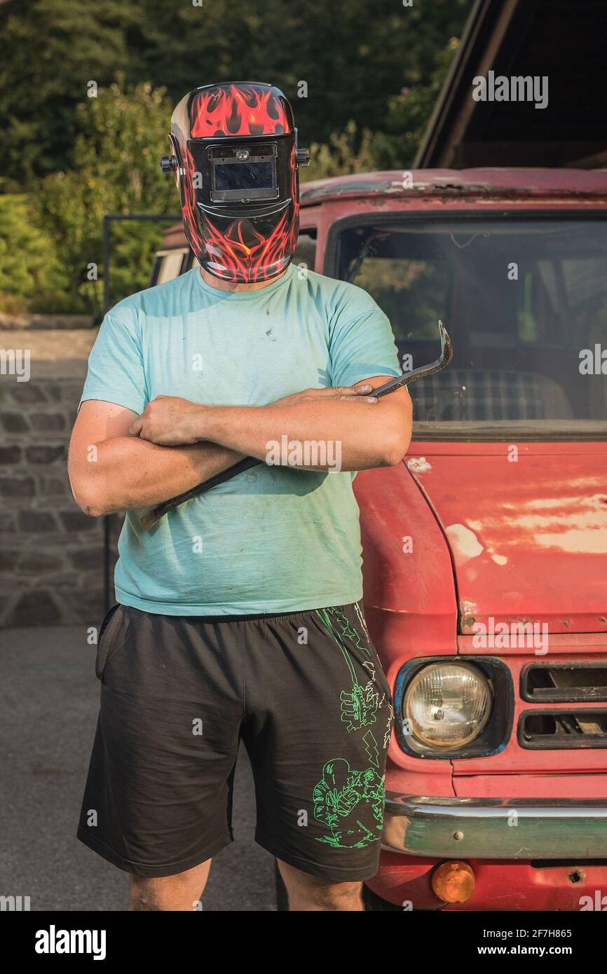 Mann mit Schweißmaske auf dem Kopf vor einem rostigen und zerstörten Oldtimer-LKW oder -Van. Auto wird wiederhergestellt. Harter Kerl, der ein Auto wieder herstellt Stockfoto