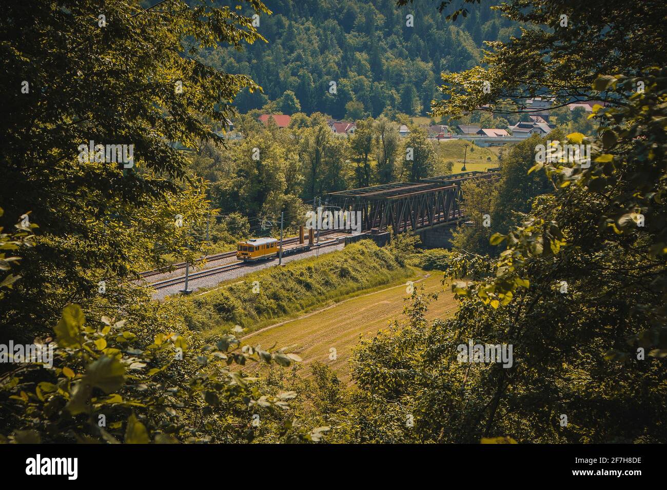 Gelber Arbeitszug mit Draisine fährt auf einer zweigleisigen Eisenbahnlinie weg von einer Metallbrücke. Romantischer Blick durch ein Loch im Laub. Stockfoto