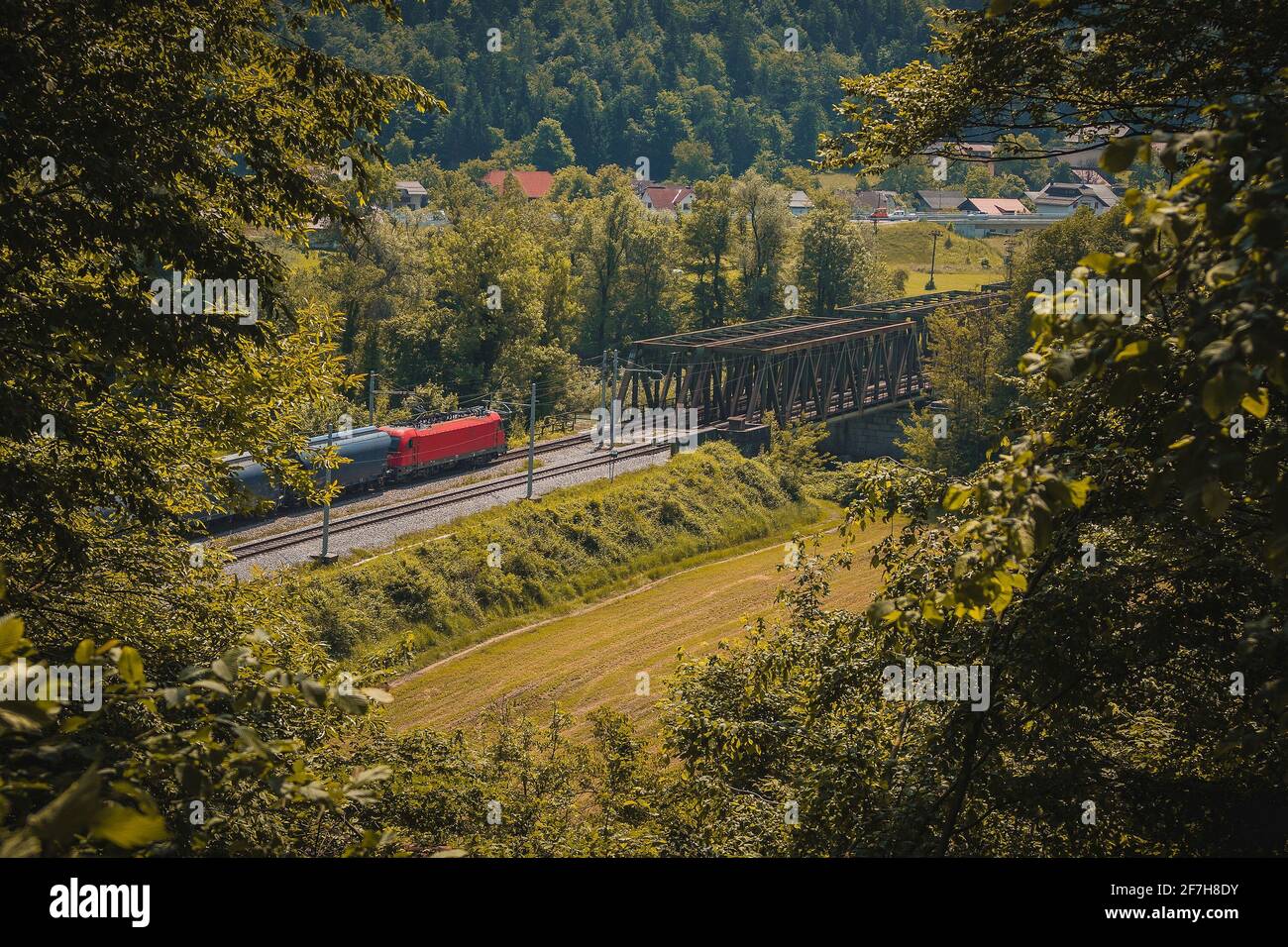 Der europäische Güterzug mit roter Lokomotive fährt auf einer zweigleisigen Eisenbahnlinie in Richtung einer Metallbrücke. Romantischer Blick durch ein Loch in Foli Stockfoto