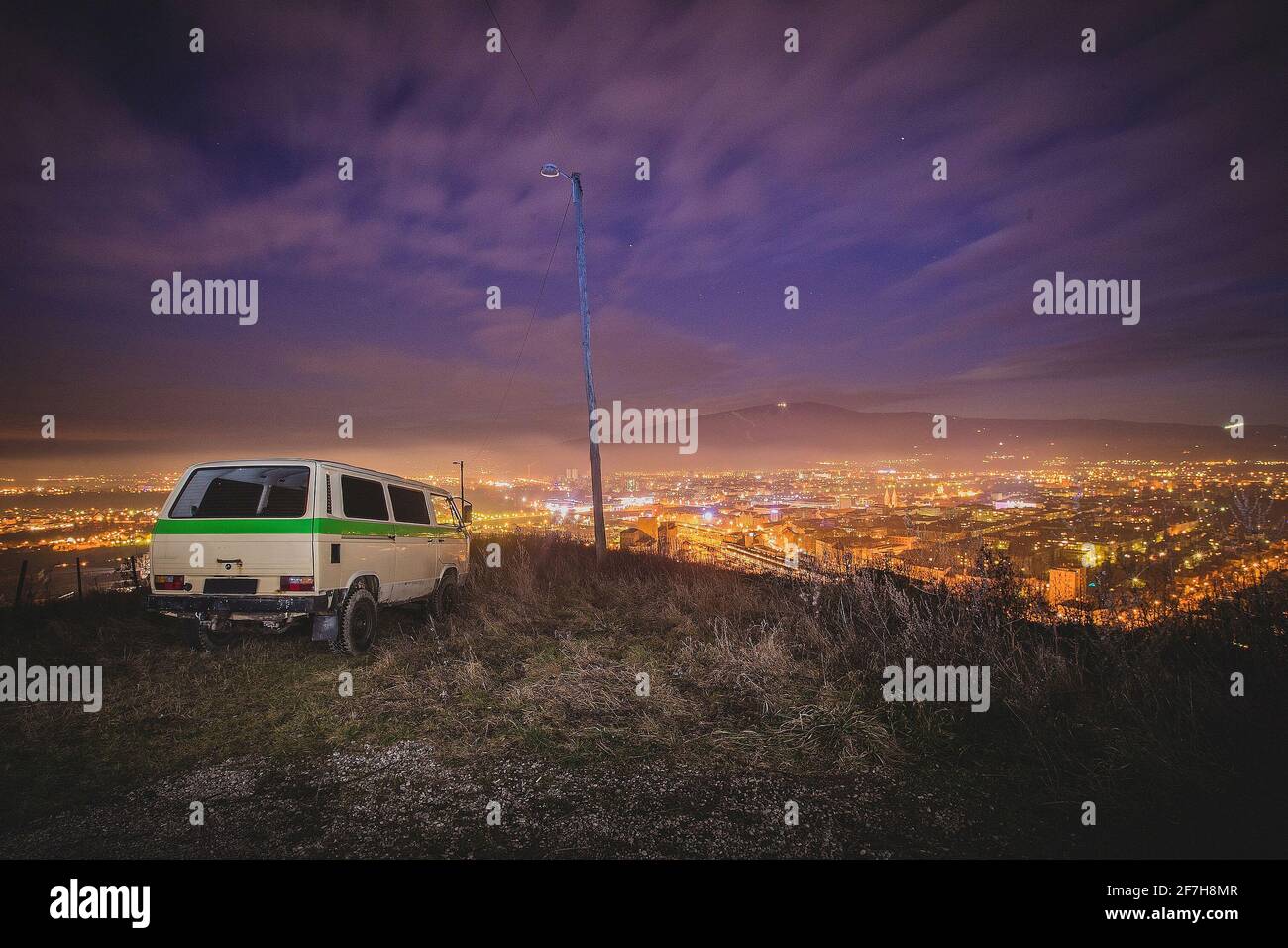 Nachtpanorama der Stadt Maribor mit einem auf dem Hügel geparkten Van. Urbane Flucht mit einem Retro-Van, mit Blick auf die Stadt in der Nacht. Stockfoto