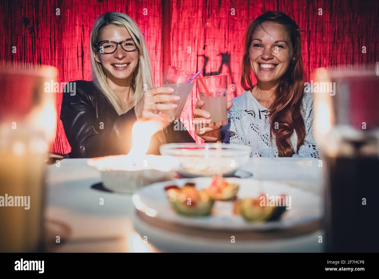 Zwei junge Frauen, eine Blondine und ein Rotschopf, jubeln am frühen Abend  mit Gläsern voller Gin Tonic Drink und Trinkhalmen auf einem Gartenpary mit  Kerze Stockfotografie - Alamy