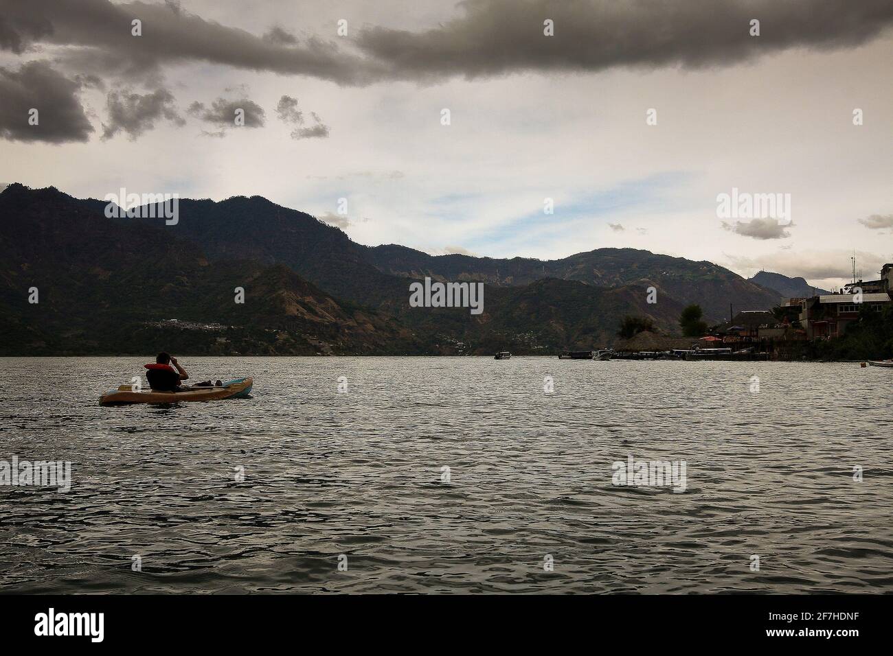 Eine Person im Kajak, Kajak auf den Gewässern des Atitlan Lake in Guatemala. Wolkiger Tag mit Bergen im Hintergrund und San Pedro Dorf. Stockfoto