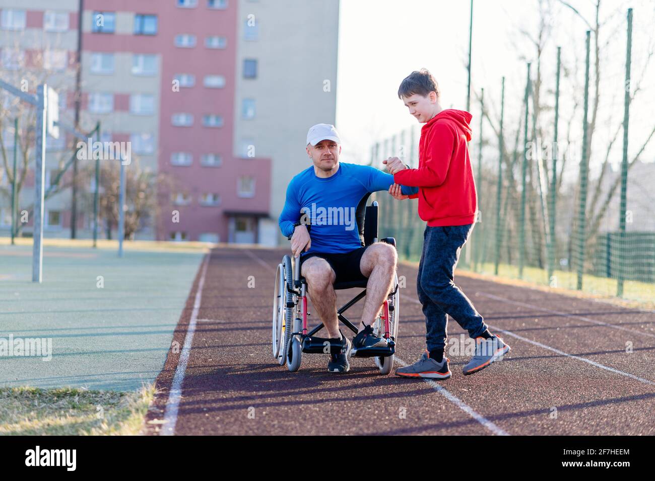Ein Teenager-Sohn hilft seinem Vater, auf einer Leichtathletik-Strecke aus seinem Rollstuhl aufzustehen. Stockfoto