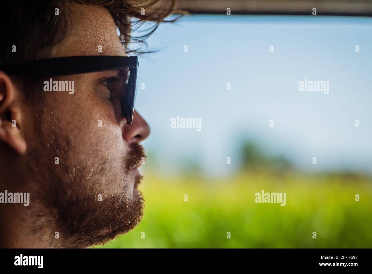 Seiten- oder Profilbild eines jungen Mannes, der mit offenem Fenster ein Auto fährt und die grünen Felder und den blauen Himmel freilegt. Vintage-Effekt-Bild mit ret Stockfoto