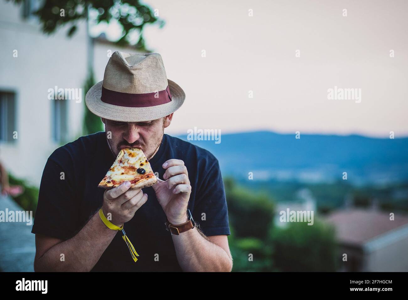 Hipster aussehender Mann mit Strohhut, der abends eine Pizza hält und mit einer französischen Landschaft im Hintergrund isst. Stockfoto