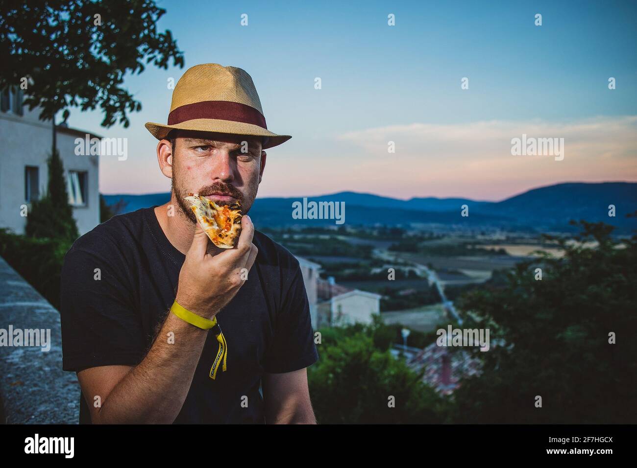 Hipster aussehender Mann mit Strohhut, der abends eine Pizza hält und mit einer französischen Landschaft im Hintergrund isst. Stockfoto