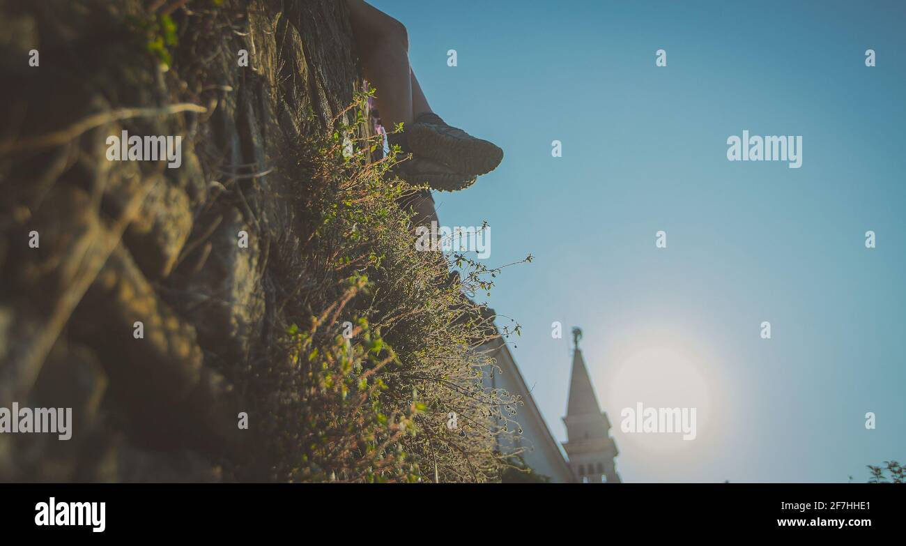 Beine einer unbekannten Frau, die an einer Backsteinmauer mit starkem Morgenlicht und Kirchturm im Hintergrund hängen. Stockfoto