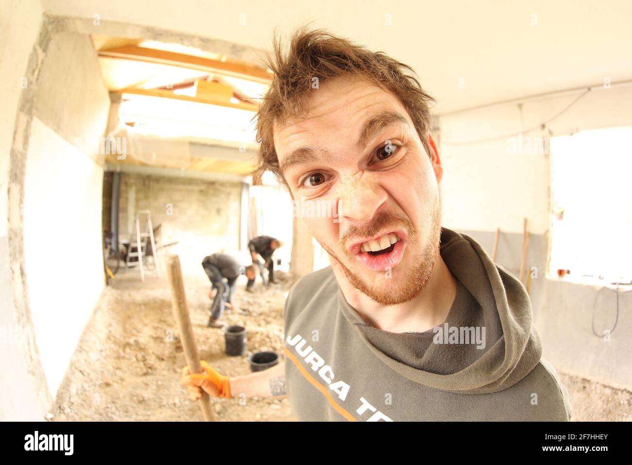 Ein Arbeiter, der ein dummes Gesicht beim Renovieren eines Hauses macht. Andere Arbeiter im Hintergrund. Fisheye-Ansicht. Stockfoto