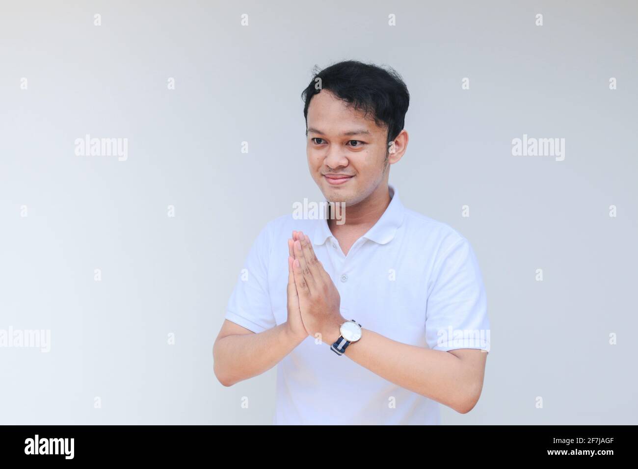 Junger asiatischer Mann in weißem Hemd geben Namaste Grußhände. Indonesischer Mann auf grauem Hintergrund. Stockfoto