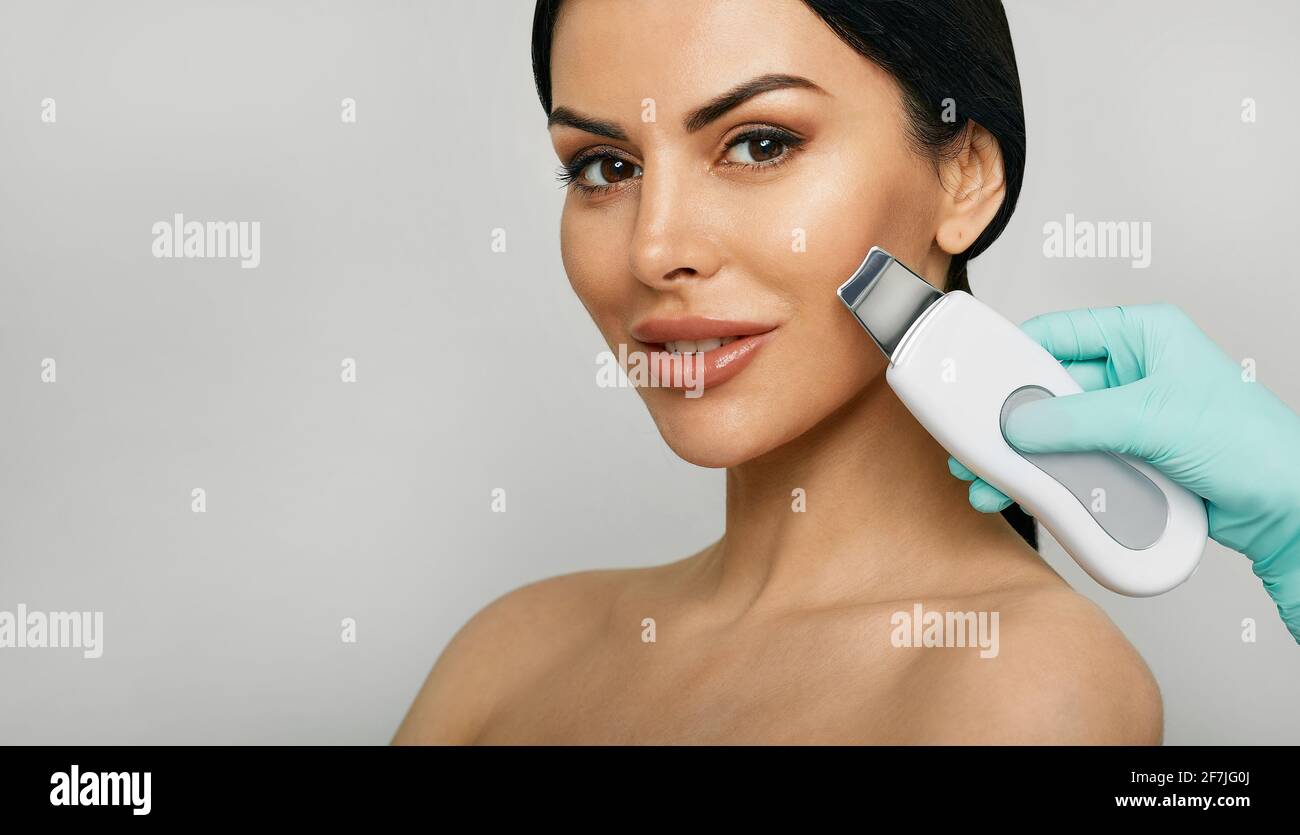 Ultraschallpeeling im Gesicht. Gemischtes Frauengesicht während des Ultraschallschälens der Haut auf grauem Hintergrund. Reinigende Gesichtshaut Stockfoto
