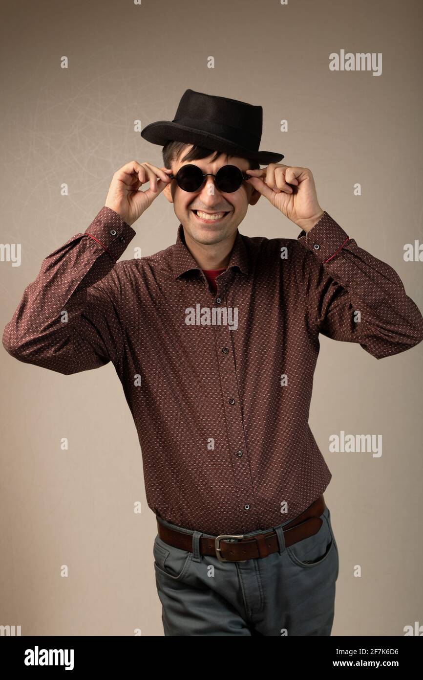 Brutaler Macho-Mann mit Hut und runder Sonnenbrille, isoliert auf dem Hintergrund des Studio-Kopierraums stehend. Trendige Mode junge kaukasischen Kerl lächeln berührend ey Stockfoto