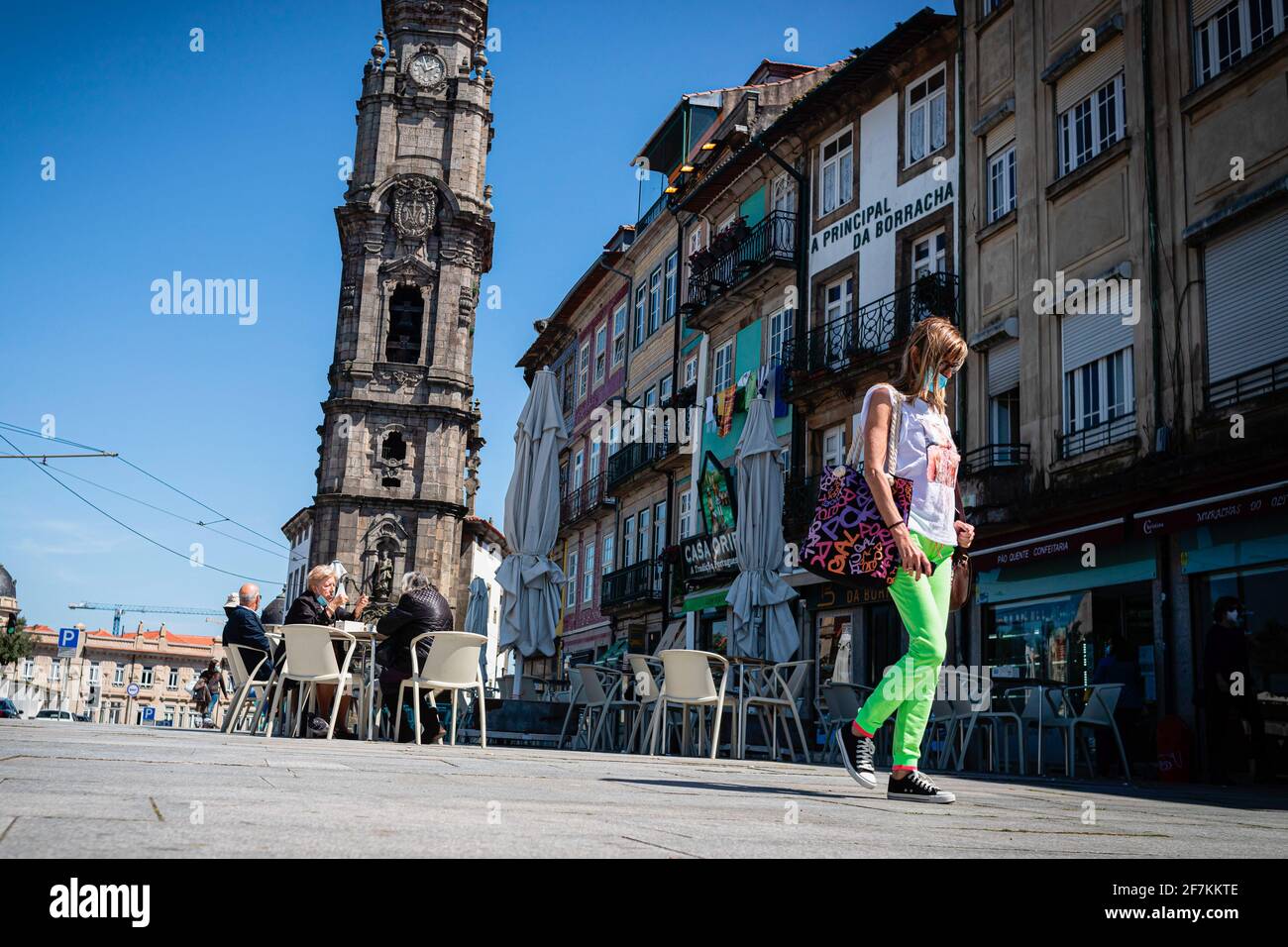 Porto, Portugal. April 2021. Eine Frau mit einer Gesichtsmaske geht zu Fuß, während im Hintergrund Menschen auf einer Esplanade direkt vor dem Clerigos Tower sitzen gesehen werden. Die zweite Phase des Definationsprozesses in Portugal begann am 5. April. Restaurants und Cafés durften Menschen auf den Tischen im Freien in Gruppen von 4 Personen pro Tisch beherbergen und die Menschen können nun mit mehr Freiheit durch die Stadt gehen. Kredit: SOPA Images Limited/Alamy Live Nachrichten Stockfoto