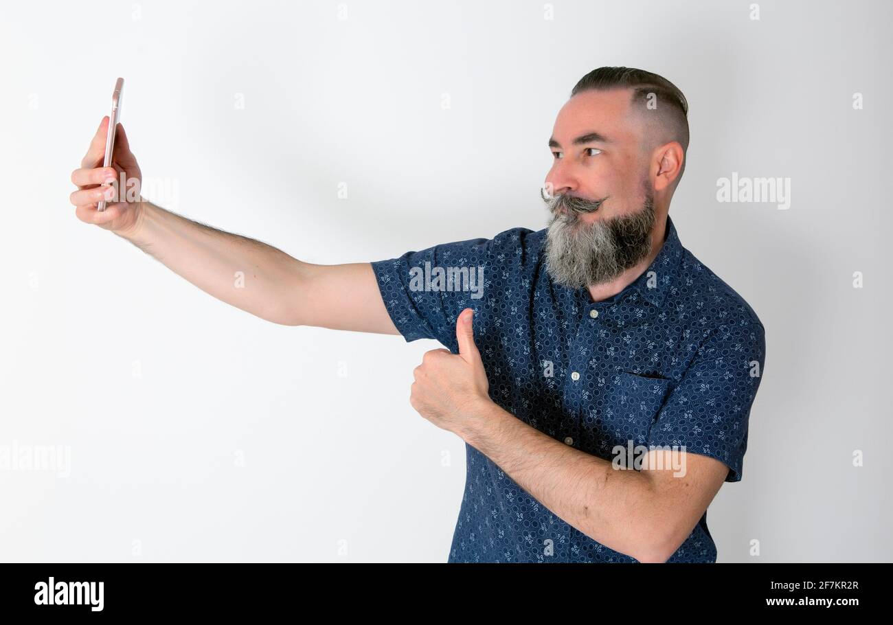 Hipster mit einem halbweißen Bart von 40-45 Jahren kaukasisch Eine Geste zur Genehmigung seines Smartphones machen, während er es hält Mit der anderen Hand und Stockfoto
