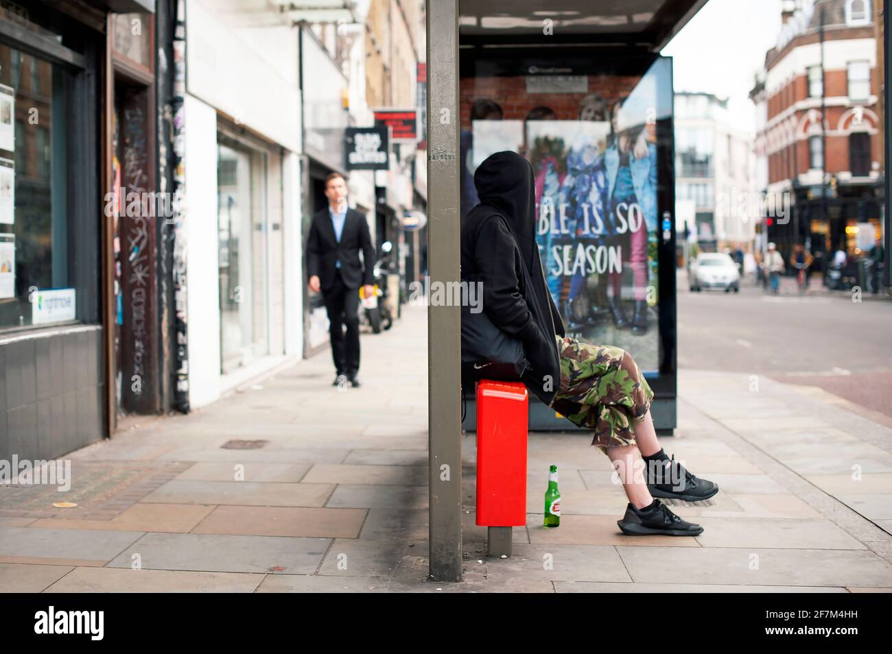 Junger Mann im Kapuzenpullover sitzt in der Bushaltestelle und wartet auf den Bus. Alltag in der Stadt. Bethnal Green Road, East London, Großbritannien. Juli 2015 Stockfoto