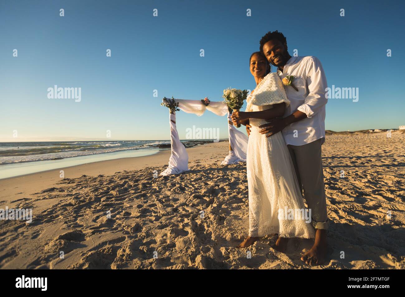 afroamerikanisches Paar, das verliebt heiratet und sich am Strand umarmt und vor die Kamera schaut Stockfoto