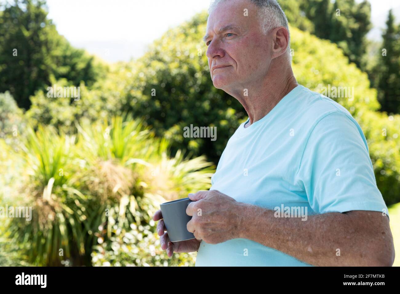 Kaukasischer älterer Mann, der im sonnigen Garten steht, die Kaffeetasse hält und wegschaut Stockfoto