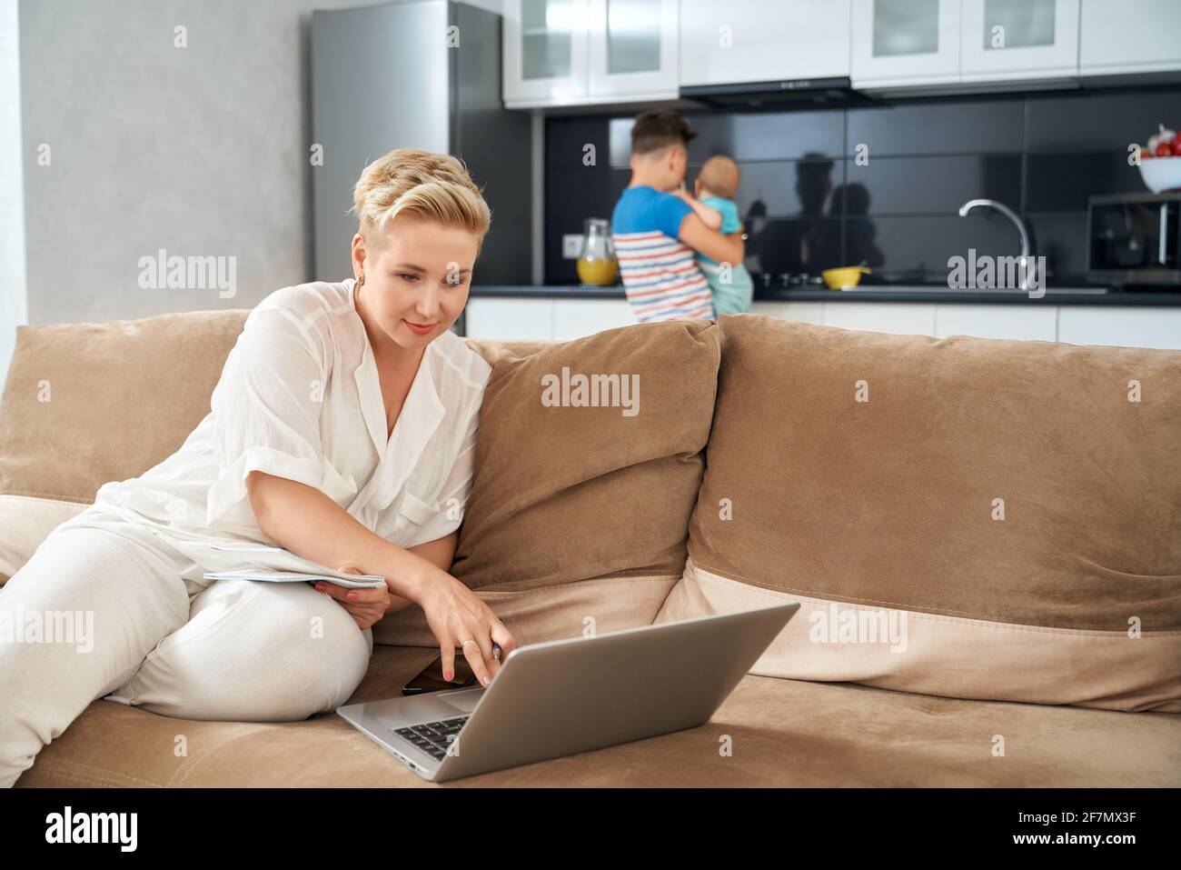 Attraktive Frau, die auf der Couch sitzt und am Laptop arbeitet, während ihr ältester Sohn sich um das kleine Baby kümmert. Fernarbeit während des Mutterschaftsurlaubs. Stockfoto