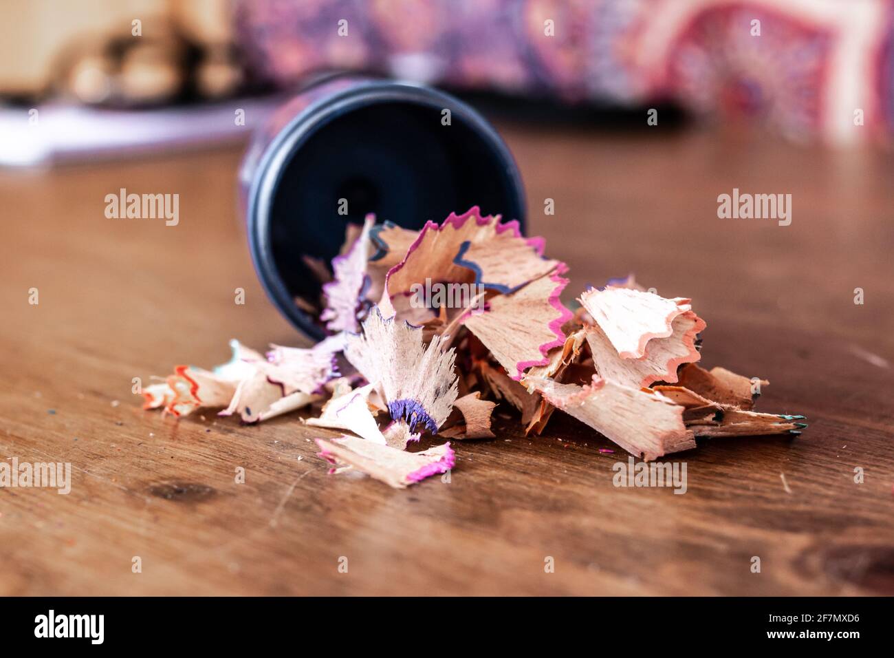 Buntstiftspangen, die aus einem blauen Bleistiftspitzer ohne Deckel auf einen Hartholztisch auslaufen, Farben sind violett, orange, rosa, gelb, Weichfokus. Stockfoto