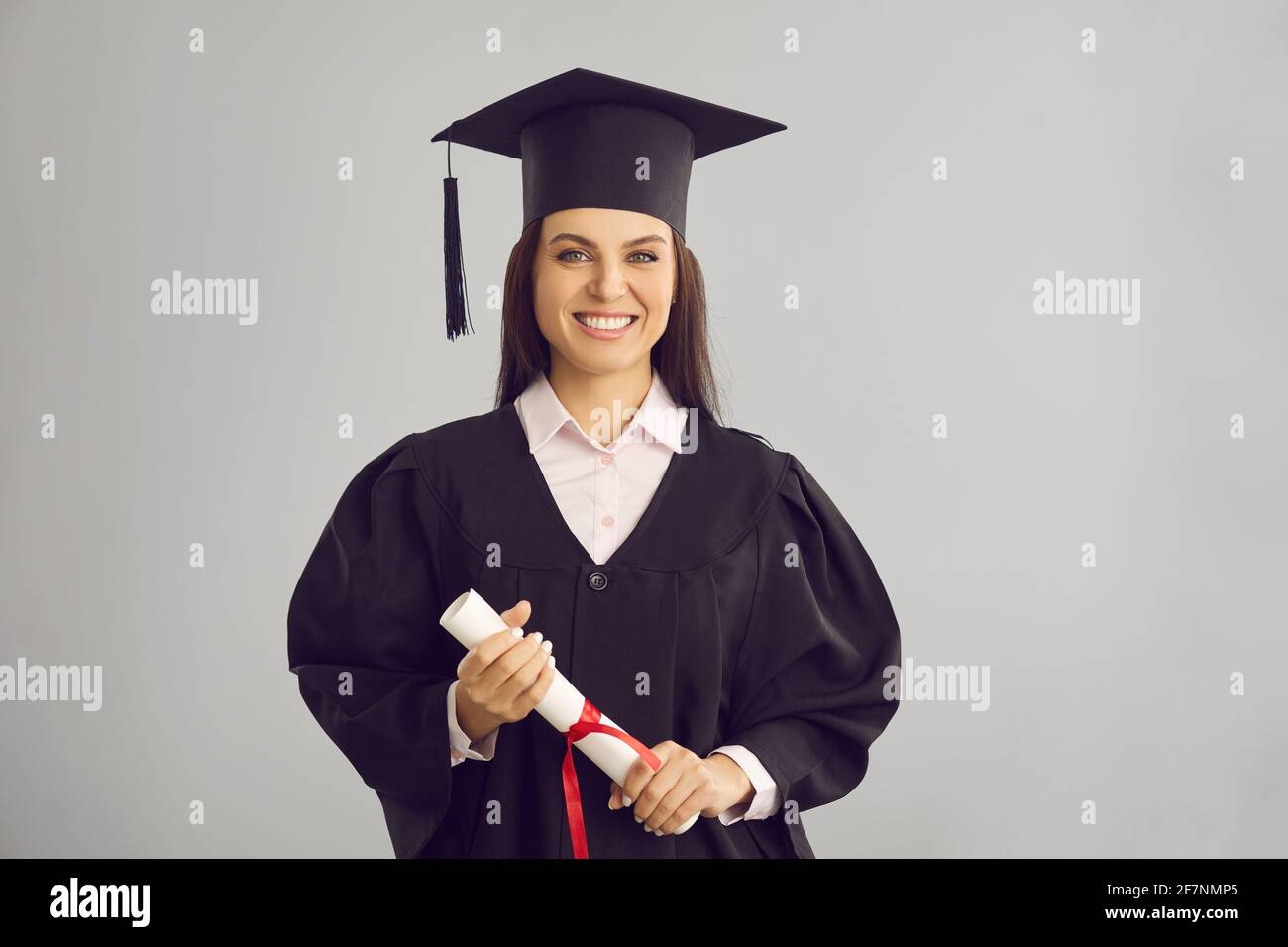 Weibliche Absolventin, die auf einem grauen Hintergrund mit einem Diplom in den Händen steht und lächelt. Stockfoto