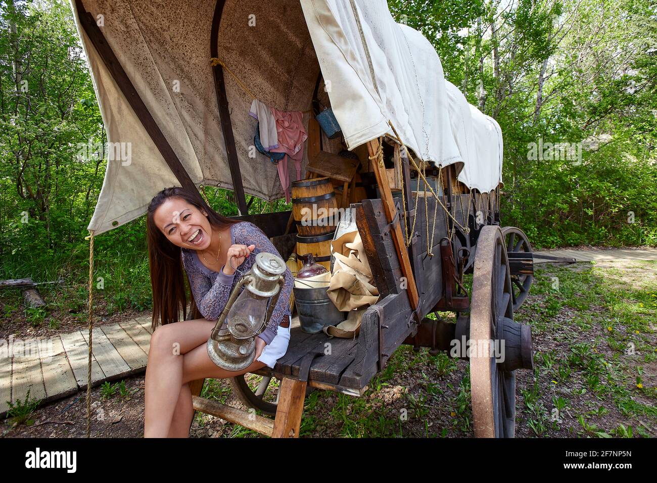 Aufgeregte asiatische Frau mit Retro-Öllampe, die die Kamera anschaut und lächelt, während sie auf einem alten Wagen sitzt, während sie in Fort Edmonton in Alberta, Kanada, besucht Stockfoto