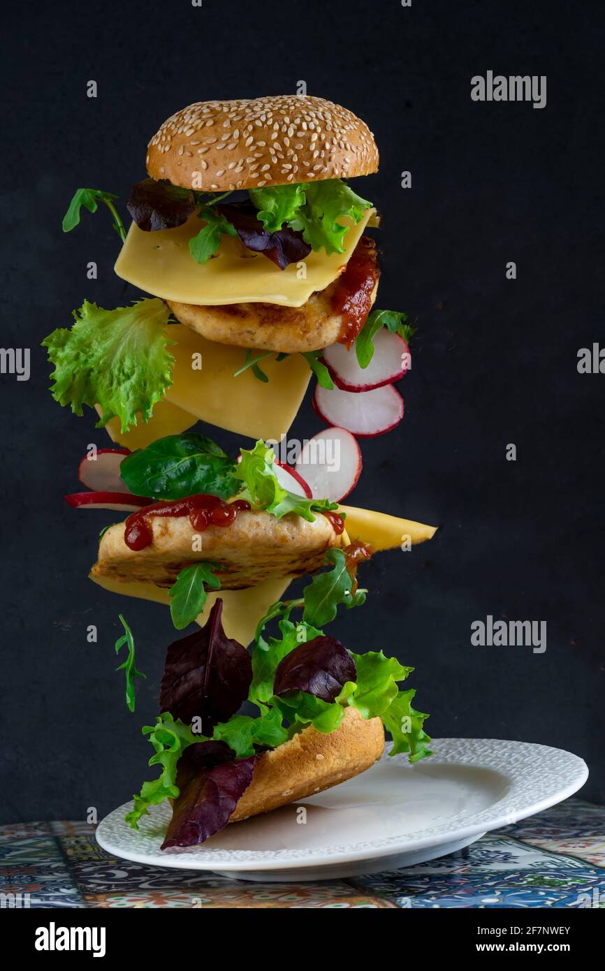 Schwebender Burger. Fliegende Nahrung. Hamburger-Brötchen, doppeltes Hühnerschnitzel mit Käse, Salat, Rucola und Rettich. Fleisch, Gemüse und Brot. Zutaten auf schwarzem Hintergrund. Stockfoto