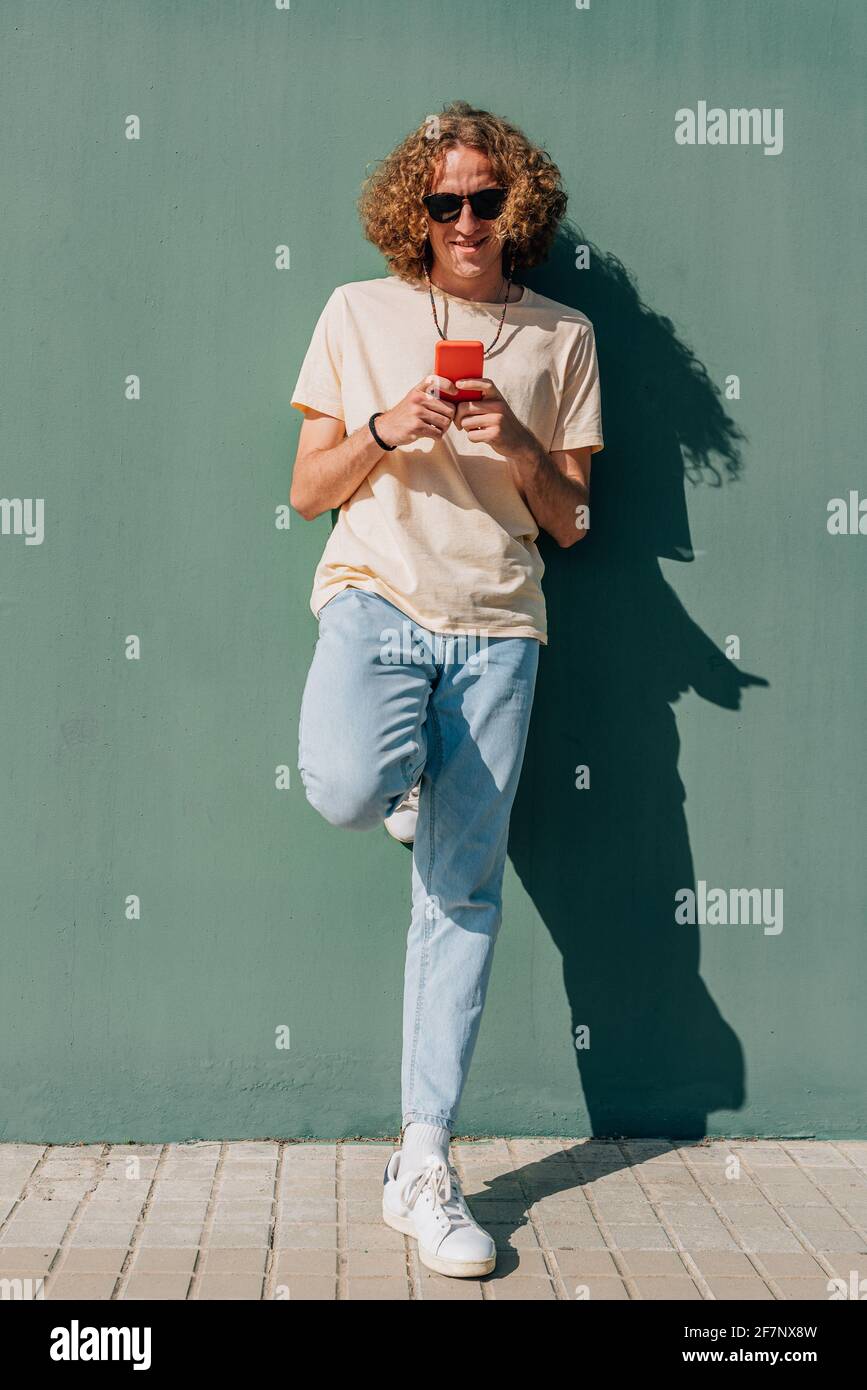 Vertikales Ganzkörperporträt eines jungen kaukasischen Mannes mit einem roten Smartphone. Er ruht auf einer grünen Mauer und lächelt. Sonniger Tag, der tiefe Schatten projiziert Stockfoto