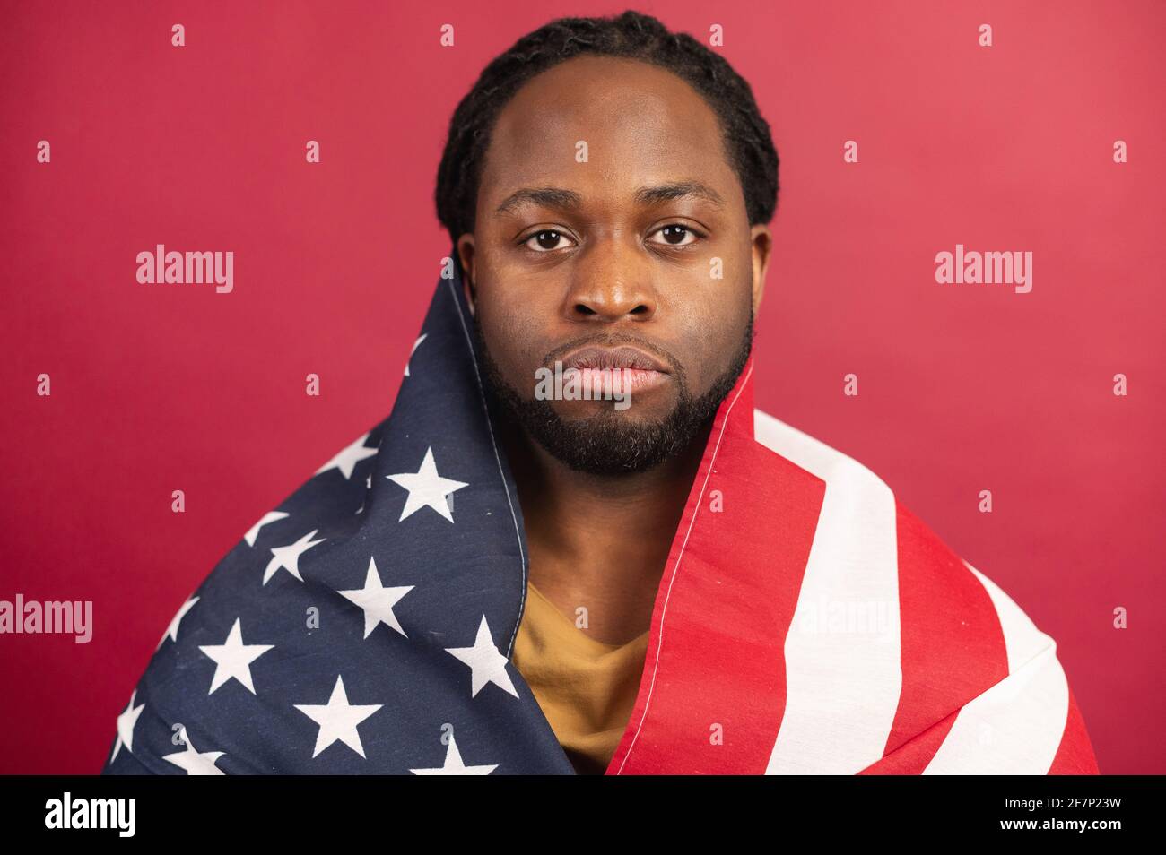 Close Portrait of serious trauriges hurted dark skinned american bärded man covered with usa flag, Head shot, looking at camera, isolated over Red background, depends his equal rights, lives in Freedom Stockfoto