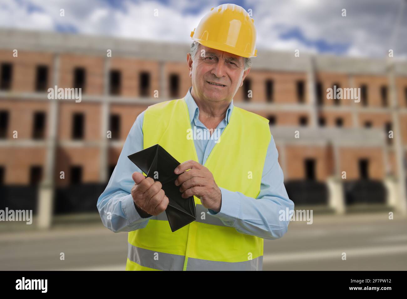 Arme alte männliche Baumeister trägt Hardhat und fluoreszierende Weste zeigen Leeres Leder-Portemonnaie mit Baugebäude im Hintergrund Stockfoto