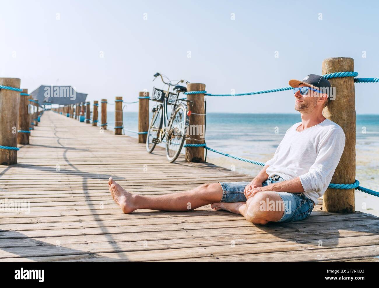 Porträt eines glücklichen lächelnden Mannes in leichten Sommerkleidung und Sonnenbrille sitzen und genießen Zeit auf hölzernen Seebrücke. Sorgloser Urlaub in Tropf Stockfoto