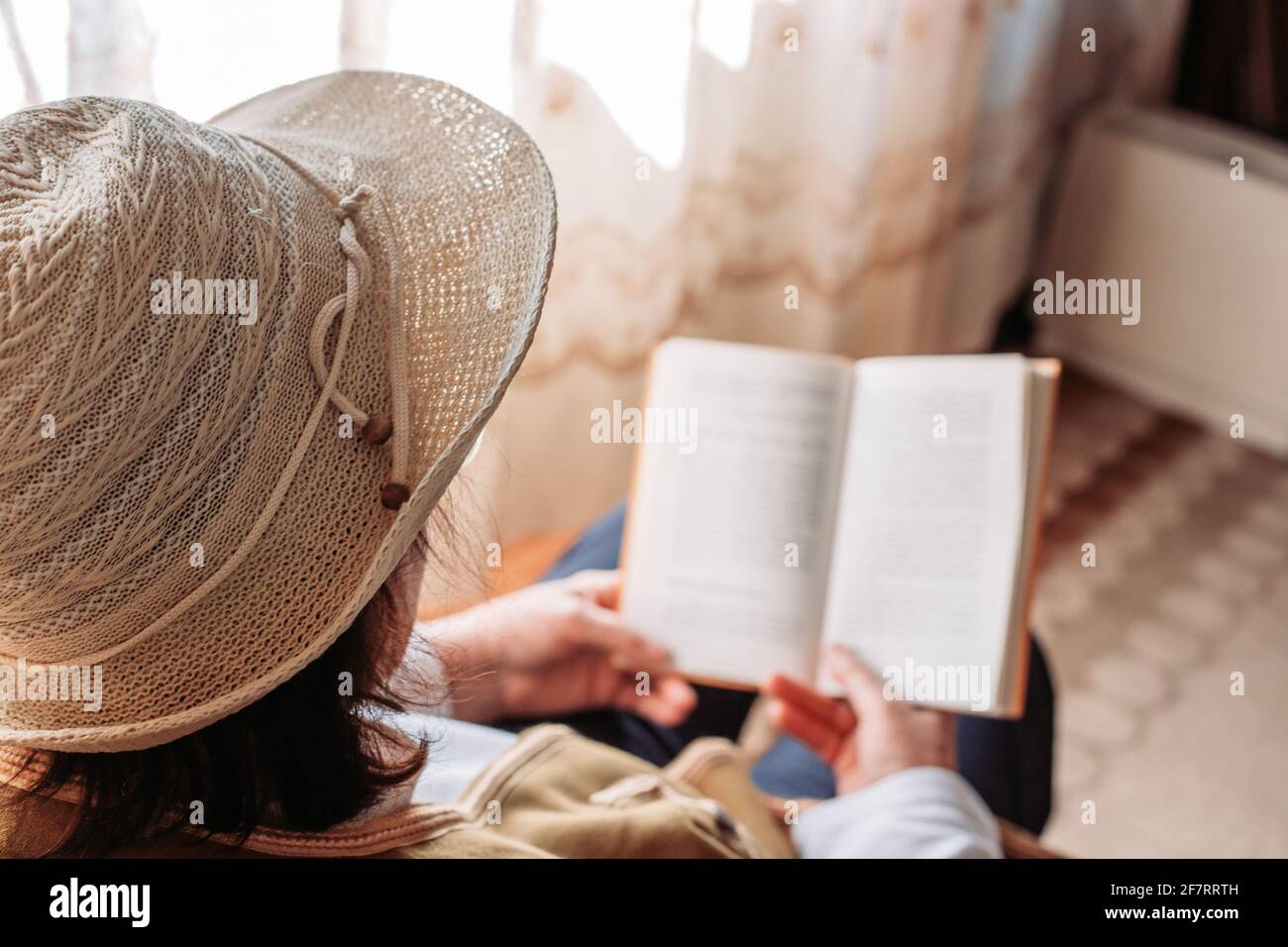 Ältere Frau mit Hut auf, liest ein Buch am Fenster. Nachmittagslicht Stockfoto