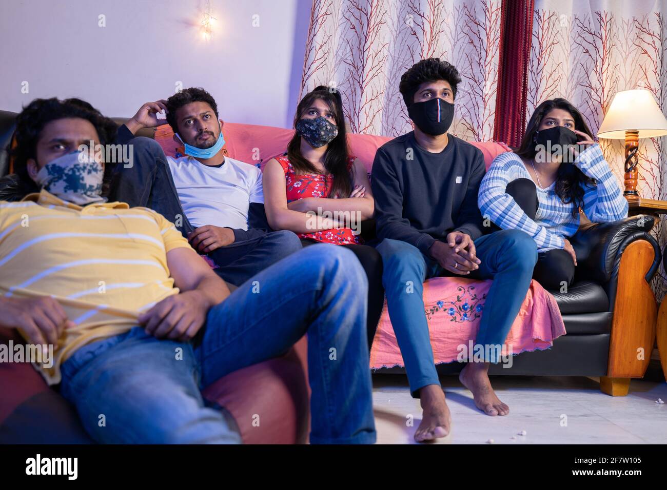 Eine Gruppe von Freunden mit medizinischer Gesichtsmaske ist damit beschäftigt, einen Film zu sehen Oder Online-Webs-Serien, die während des Studiums auf dem Sofa sitzen Urlaub wegen Coronaviru covid Stockfoto