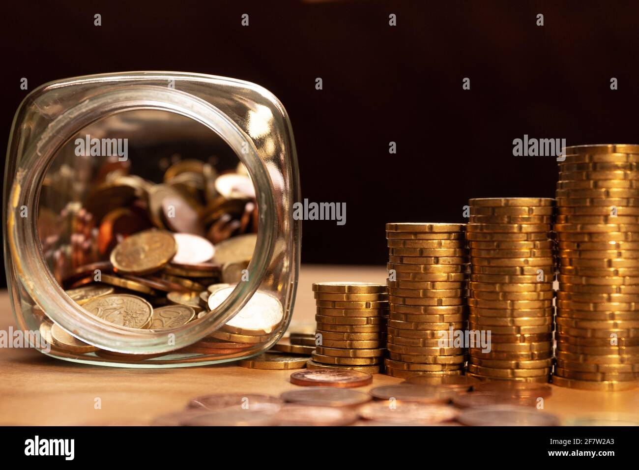 Ein halb leeres Glas mit Münzen mit Münzstapeln und mehr Münzen auf einem Tisch. Sparen Sie Geld Konzept Banner Stockfoto