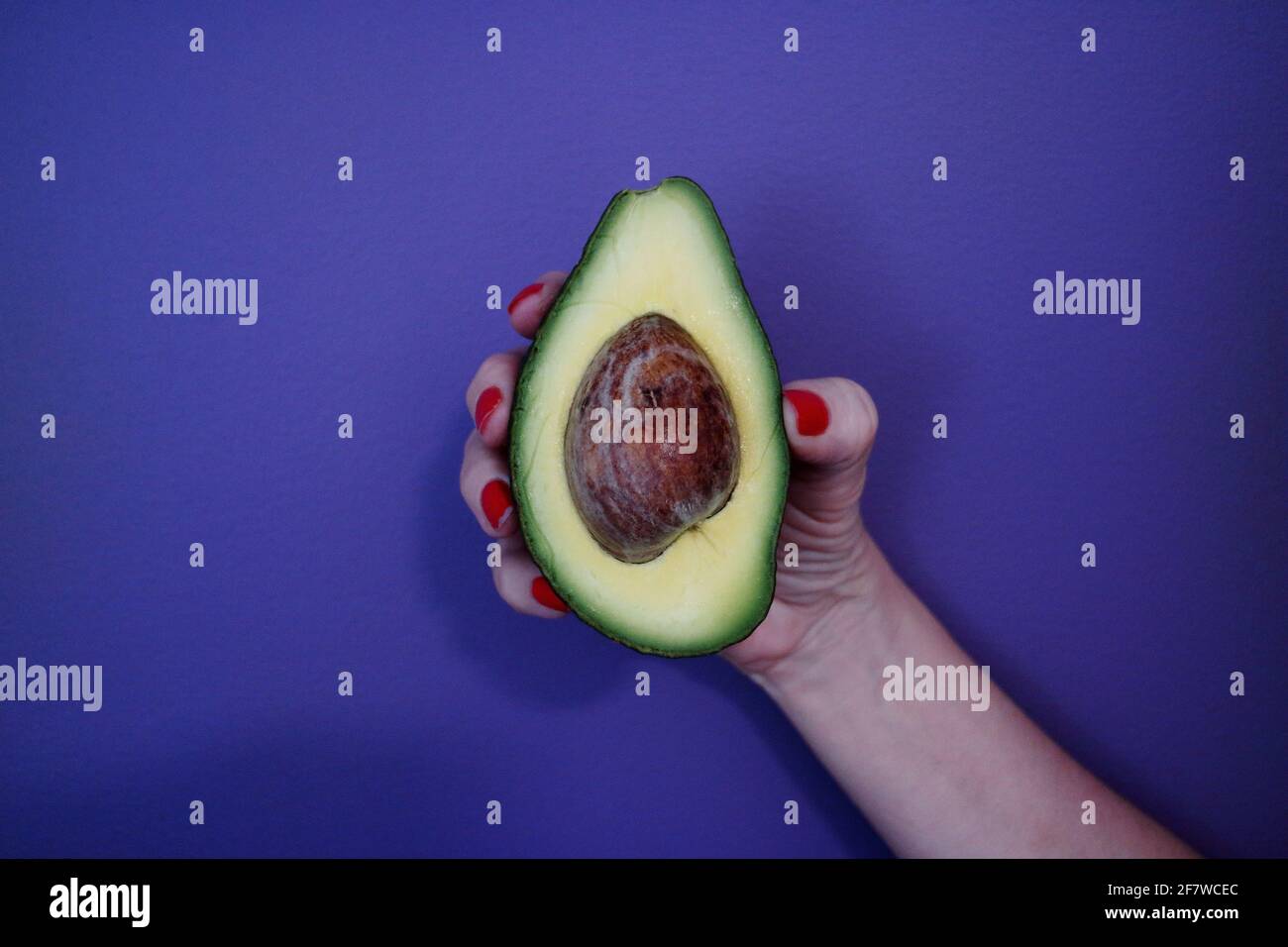 Nahaufnahme der Hand einer Frau mit rot lackiertem Finger Nägel halten eine halbgeschnittene Avocado gegen den violetten Hintergrund Stockfoto