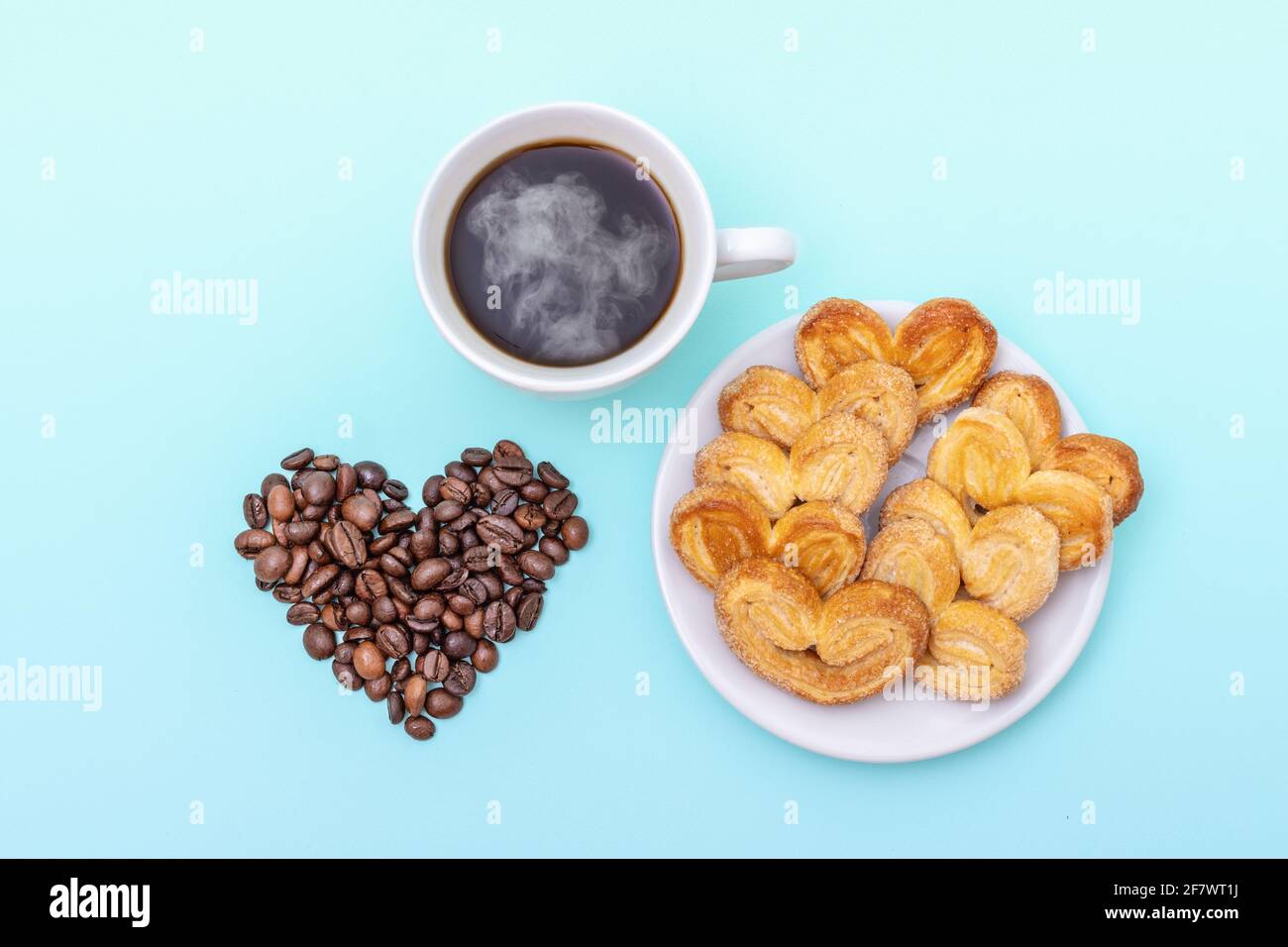 Dampfende Kaffeetasse, herzförmige Kekse, herzförmige Kaffeebohnen auf blauem Hintergrund, Draufsicht. Frühstück am Morgen. Frühstück für die Liebsten. Stockfoto