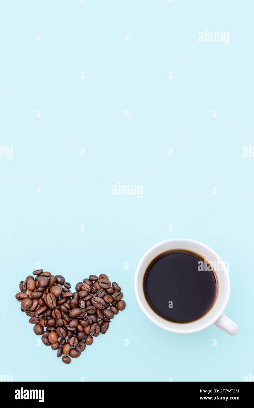 Weiße Tasse schwarzen Kaffee und Körner von Kaffee in Form eines Herzens auf einem blauen Hintergrund, Kopierraum, Draufsicht, vertikale Form. Kaffee am Morgen Stockfoto