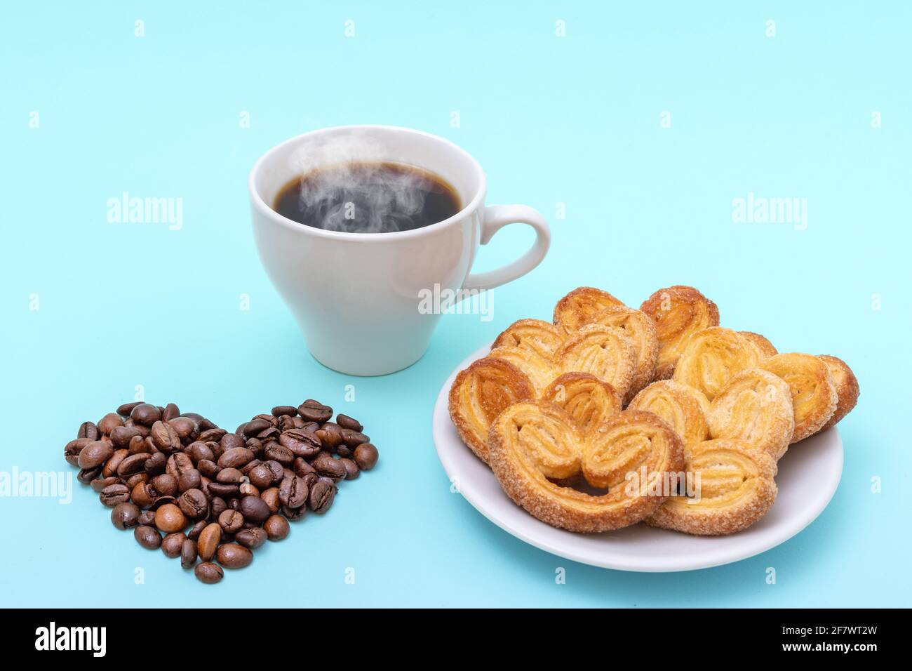 Dampfende Kaffeetasse, herzförmige Kekse, herzförmige Kaffeebohnen auf blauem Hintergrund. Frühstück am Morgen. Frühstück für die Liebsten. Stockfoto